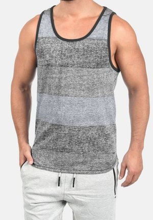 Man draagt een mouwloos grijs tanktop met horizontale strepen en lichtgrijze joggingbroek, staat met één hand in zijn zak.
