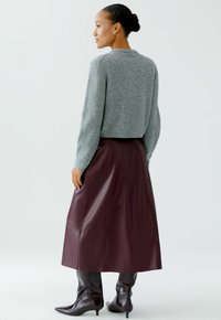 Vrouw in een grijze sweater, bordeauxrode leren midi-rok en zwarte puntige enkellaarsjes, zijwaarts staand op een effen lichte achtergrond.