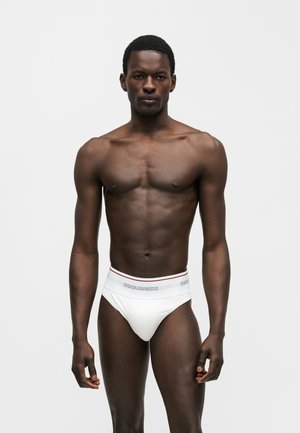 Αθλητικός άνδρας με σκουρόχρωμο δέρμα στέκεται κοιτώντας εμπρός, φοράει λευκά εσώρουχα DSQUARED2 σε απλό ανοιχτό φόντο.