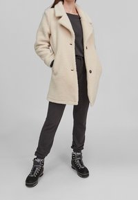 Abrigo de forro polar crema con solapas en pico, dos bolsillos frontales y botones negros, llevado sobre un conjunto gris oscuro y combinado con botas de senderismo negras.