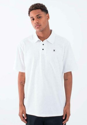 Camisa polo blanca con una tapeta de dos botones, mangas cortas y un pequeño logo negro en el lado izquierdo del pecho. Tejido suave y texturizado.