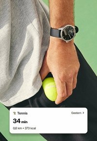 Palla da tennis nella tasca di pantaloni sportivi verde e neri, mano che stringe la palla; indossa un orologio argentato con cinturino nero e una maglietta grigia.