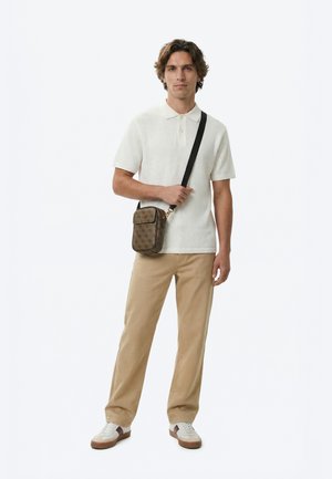 Mann iført hvit strikket polo skjorte, beige bukser, hvite og brune joggesko, bærende en brun skulderveske med svart stropp.