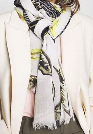 Femme portant un blazer crème sur un haut rose avec une écharpe blanche ornée de motifs abstraits noirs et jaunes enroulée autour du cou.