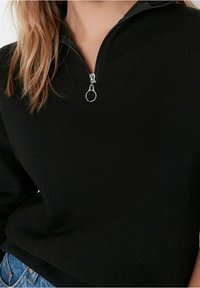 Sudadera negra con cremallera y cuello alto, hardware de cremallera plateado, textura de tela suave y corte holgado. Combinada con jeans azules.