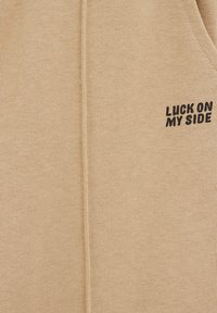 Beige huppari, jossa on ribattu pinta, jossa on musta kirjailtu teksti "LUCK ON MY SIDE". Esittelee yksinkertaista muotoilua vetoketjutta tyylissä.