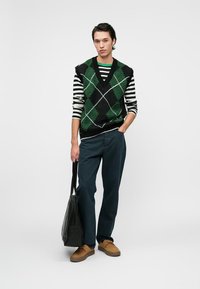 Weste mit schwarz-grünem Argyle-Muster über einem schwarz-weißen gestreiften Langarmshirt, kombiniert mit dunklen Hosen und braunen Schuhen, während eine schwarze Tasche gehalten wird.