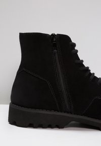 Botín de ante negro con cremallera lateral, superficie texturizada y suela resistente con patrones de relieve pronunciados.