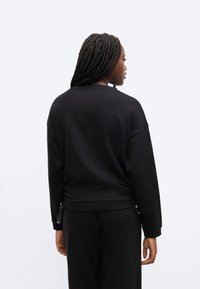 Musta, pitkähihainen collegepaita väljässä istuvuudessa, jossa on pyöreä kaula-aukko, laskeneet olkapäät ja nauhakiinnitys helmassa.