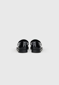 Mustat nahkakengät, joissa on litteä korko ja uurteiset reunat, omaavat sileän pinnan ja minimalistisen muotoilun, nähtynä takaa.