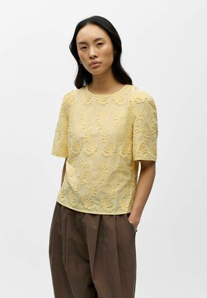Femme aux longs cheveux noirs portant un haut jaune à manches courtes brodé de fleurs et un pantalon marron ample, debout les mains dans les poches.