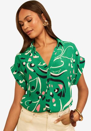 Frau trägt eine grüne Bluse mit kurzen Ärmeln und einem bunten abstrakten Muster sowie beige Hosen, accessoriert mit großen Ohrringen und Armbändern.
