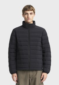 Jeune homme aux cheveux courts portant une veste matelassée noire zippée et un pantalon beige, debout devant un fond clair uni.