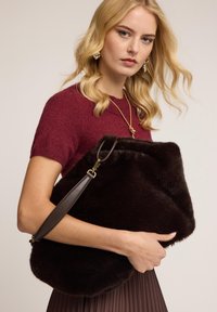 Ruskea turkishaikku, jossa on pehmeä rakenne, kulmikas muoto ja ruskea nahkaremmi. Mallilla on päällään burgundin värinen neulepaita ja laskostettu hame.