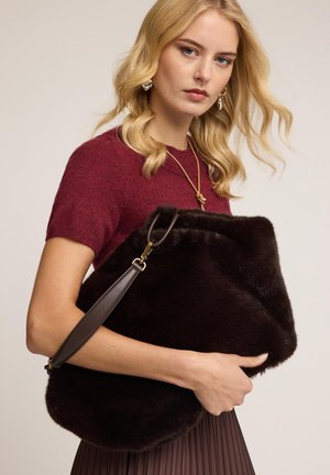 Bolso de piel marrón con una textura suave, forma angular y una correa de cuero marrón. La modelo lleva un suéter burdeos y una falda plisada.