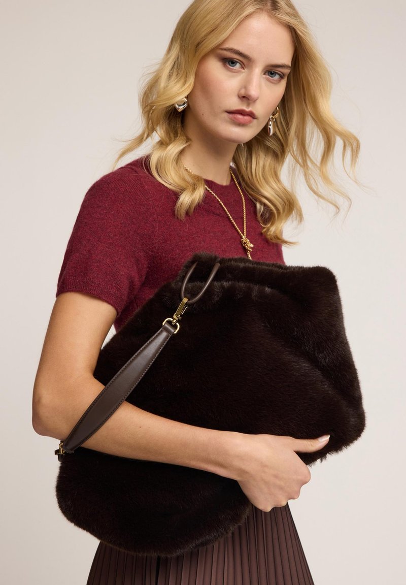 Ruskea turkishaikku, jossa on pehmeä rakenne, kulmikas muoto ja ruskea nahkaremmi. Mallilla on päällään burgundin värinen neulepaita ja laskostettu hame.