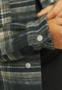 Grön och brun rutig flanellskjorta med silverfärgade tryckknappar, som visar textur och mönsterdetaljer vid manschetterna och kragen.