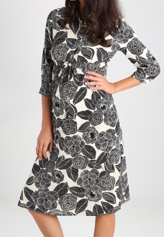 Femme portant une robe blanche longueur genou avec des motifs floraux et feuilles noirs, manches trois-quarts, et une ceinture nouée à la taille.