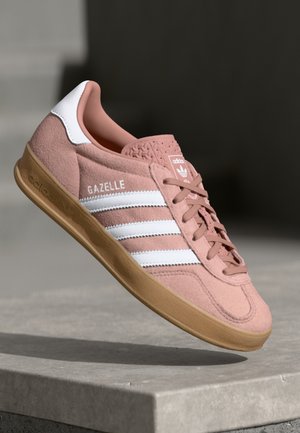 Pink Adidas Gazelle sneaker med hvide striber og gummisål, vist fra siden mod en sløret neutral baggrund.