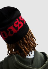 Beanie nera a maglia con testo rosso, caratterizzata da una trama di filato attorcigliato; indossata con una giacca verde oliva, mostrando una camicia bianca sotto.