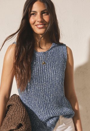 Smilende kvinde med langt mørkt hår iført en ærmeløs blå striktop, hvide bukser og en gylden halvmånehalskæde, mens hun holder en brun flettet sweater.