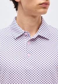 Άνδρας που φορά ροζ πουκάμισο με μικρό μπλε μοτίβο σε σχήμα καρδιάς, το γιακά ανοιχτό και τρία ορατά κουμπιά κλεισμένα.