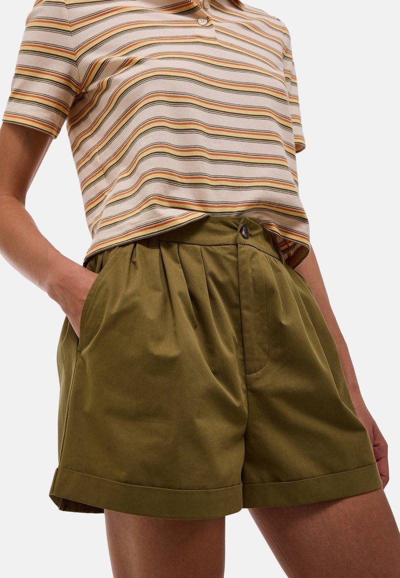 Shorts plissés vert olive avec poches latérales et ourlet replié. Associés à une chemise col beige rayée.