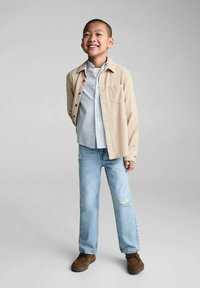 Camicia button-up beige sopra una camicia a righe blu chiaro, abbinata a jeans blu chiaro strappati e scarpe marroni. Bambino sorridente che sta in piedi con sicurezza.