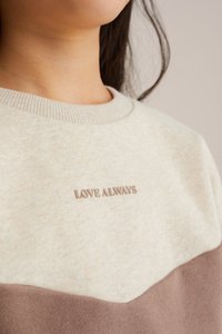 Mikina v krémovej a hnedej farbe, s rebrovaným výstrihom a vyšitým textom "LOVE ALWAYS" na hrudi. Mäkká textúra a neformálny dizajn.