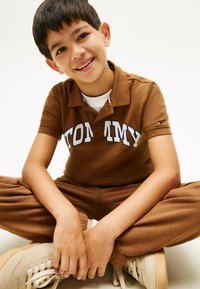 Brun polo t-shirt med hvid "TOMMY" skrift, parret med matchende brune bukser og beige sneakers. Glat tekstur, klassisk pasform.