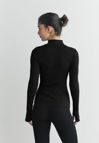 Top nero a maniche lunghe con coste, collo alto, design aderente e dettagli con fori per il pollice, abbinato a leggings neri. Texture liscia.