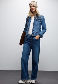 Giacca di jeans e jeans blu a vita alta abbinati a una maglietta bianca, una fascia beige e sneakers bianche, con una borsa a tracolla marrone.