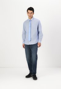 Camisa de manga larga azul claro con cuello y bolsillo, combinada con jeans oscuros y zapatillas negras. La camisa presenta un color sólido y una textura suave.