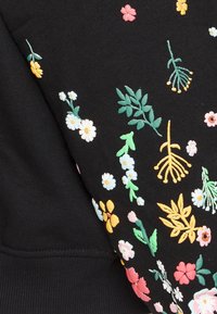 Tissu noir avec broderie florale multicolore vibrante mettant en vedette des marguerites, des feuilles et diverses fleurs en jaune, rose, blanc et vert.