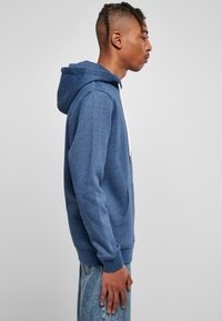 Felpa con cappuccio in materiale pile blu. Presenta una tasca frontale, lacci bianchi e polsini a costine. Vista laterale che mostra una vestibilità rilassata.