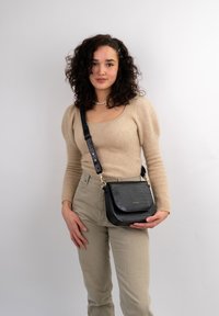 Schwarze Umhängetasche mit geprägtem Krokodilmuster, goldenen Beschlägen und verstellbarem Trageriemen. Kombiniert mit einem beigen Pullover und hellen Hosen.