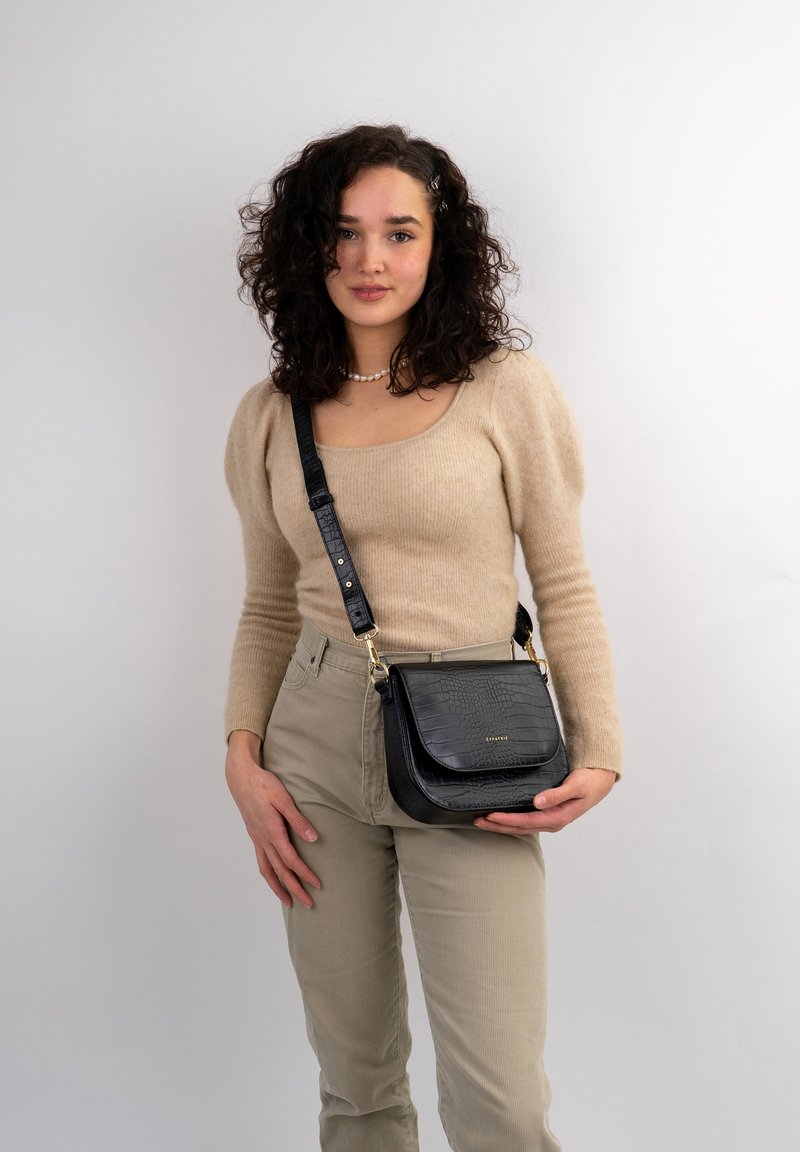Schwarze Umhängetasche mit geprägtem Krokodilmuster, goldenen Beschlägen und verstellbarem Trageriemen. Kombiniert mit einem beigen Pullover und hellen Hosen.