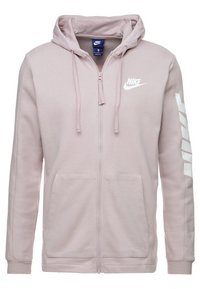 Sudadera con cremallera de color rosa claro, hecha de tejido suave. Cuenta con una capucha forrada de malla, bolsillos tipo canguro y el logo de Nike en blanco en el pecho y la manga.