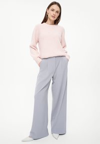 Helles rosa geripptes Strickpullover mit Perlenakzenten, kombiniert mit hellgrauen, weit geschnittenen Hosen. Das Model steht mit den Händen in den Taschen vor einem weißen Hintergrund.