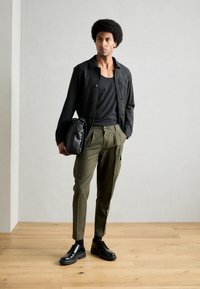 Czarna kurtka zapinana na guziki, czarny top na ramiączkach, oliwkowe spodnie cargo, czarne skórzane buty. Model trzyma czarną torbę na ramię. Tło z drewnianej podłogi.