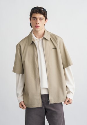 Ung mann med mørkt, krøllete hår som har på seg svart caps bak frem, beige kortermet jakke over hvit langermet skjorte, og mørke bukser mot ensfarget bakgrunn.