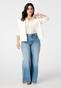 Crèmekleurige gebreide vest over een kanten blouse met knopen, gecombineerd met lichtblauwe hoge taille flare jeans. Opvallende kanten textuur en relaxte pasvorm.