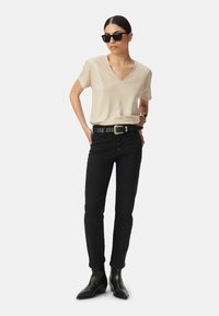 T-shirt beige a V, jeans neri a vita alta con bottoni, cintura accentuata con borchie argentate e stivaletti neri a punta.