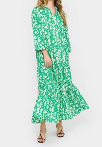 Grön blommig maxiklänning med V-ringning, trekvartslånga ärmar och en lager-på-lager volangslut. Tillverkad av lätt material med vita blommönster.