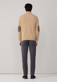 Uomo che indossa un maglione beige con toppe marroni sui gomiti, pantaloni grigio scuro e scarpe nere, in piedi di fronte a un muro grigio uniforme.