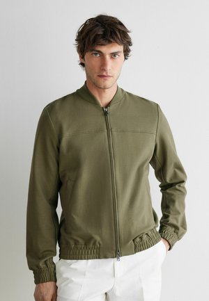 Mann trägt olivgrüne Jacke mit Reißverschluss und weiße Hose, steht vor einem schlichten hellen Hintergrund mit einer Hand in der Tasche.