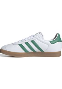Weißer Sportschuh mit grünem Drei-Streifen-Design, glattem Synthetik-Obermaterial, Schnürverschluss und Gummisohle.