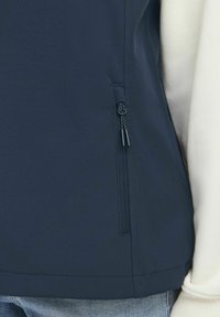 Mornarska softshell jakna s bočnom džepom na zatvarač, s glatkom teksturom i uklopljenim dizajnom, u kombinaciji s kontrastnim svijetlim puloverom.