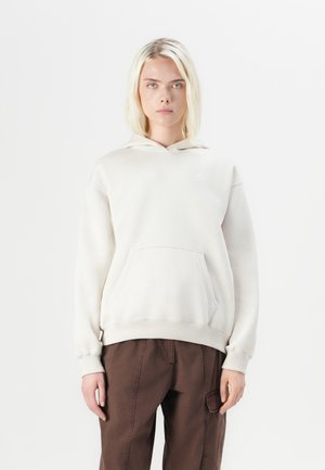 A model with shoulder-length blonde hair wears a light beige hoodie with a front pocket and brown pants, set against a plain white background.