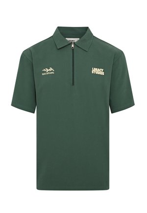 Polo shirt vert foncé, manches courtes, col zippé, avec "LEGACY STUDIOS" en jaune clair et un design graphique au-dessus du texte.
